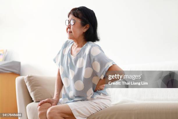 senior asian woman suffering from backache, sitting on sofa at home - heup stockfoto's en -beelden