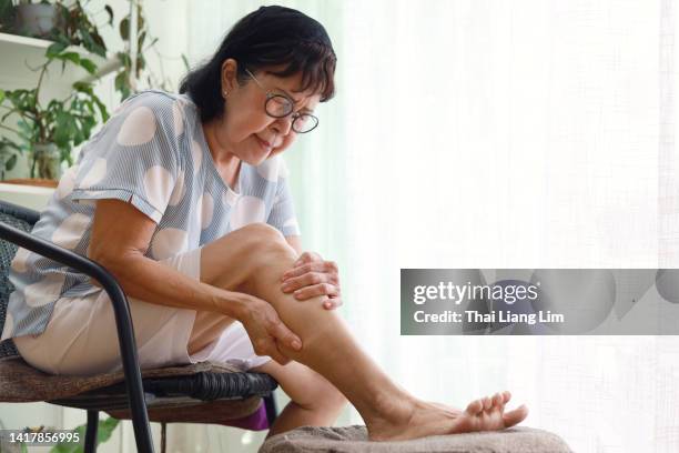 una mujer asiática mayor masajeando su pierna, sufriendo de dolor en la pierna, sentada en el sillón en la sala de estar - woman holding legs fotografías e imágenes de stock