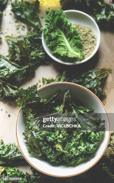 close up of healthy green kale chips in bowl on kitchen table with ingredients - cabbage family fotografías e imágenes de stock