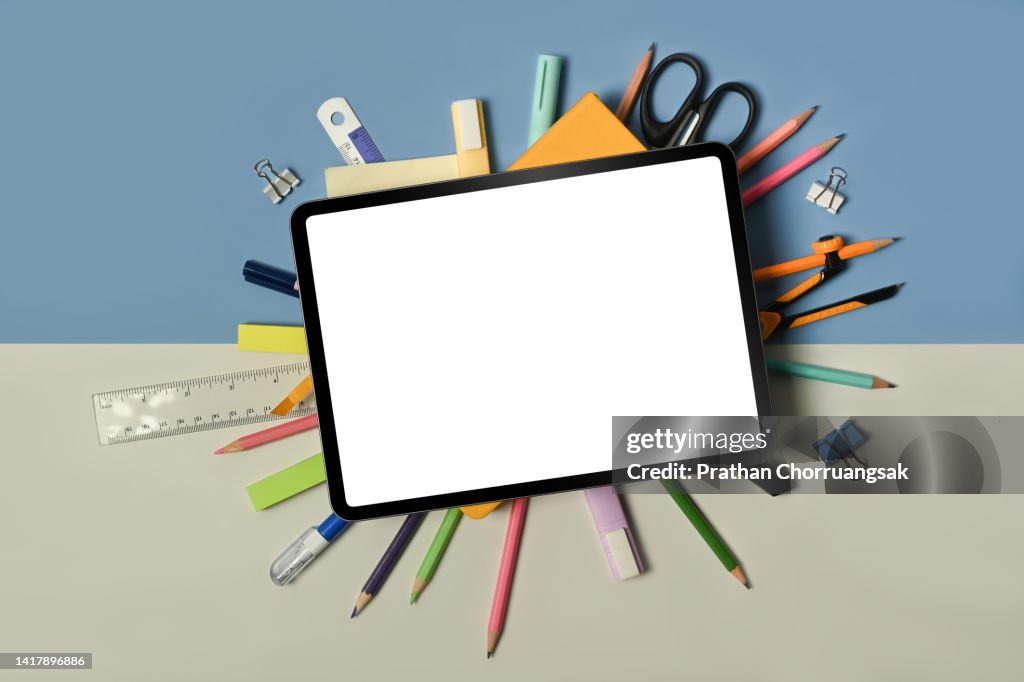Digital tablet with empty screen and school stationery bag pack on blue and beige background.