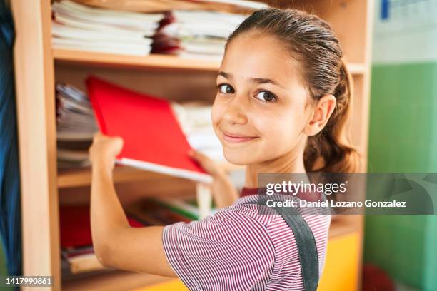 student girl looking at camera and smiling while putting textbook on shelf at school. - putting book on shelf stock pictures, royalty-free photos & images