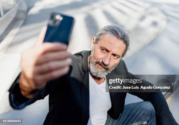 front view of a middle-aged businessman with ponytail and blazer takes a selfie with his smartphone in the city - showing off stock-fotos und bilder
