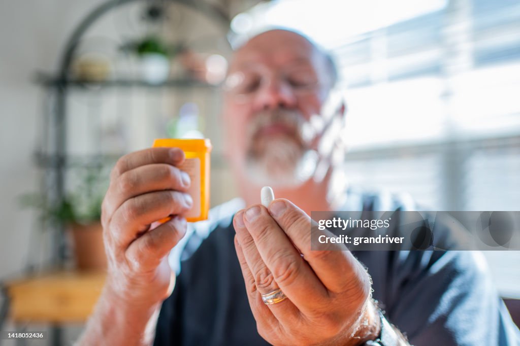 Uomo maturo che esamina i suoi farmaci di prescrizione tenendo una pillola in una mano e la bottiglia nell'altra in una casa moderna