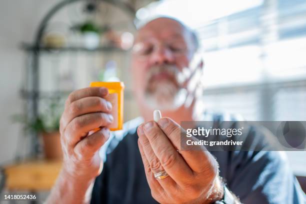 mature man scrutinizing his perscription medications holding a pill in one hand and the bottle in the other in a modern home - prescription imagens e fotografias de stock