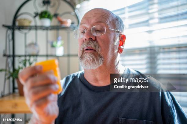 mature man scrutinizing his perscription medications holding a pill in one hand and the bottle in the other in a modern home - holding bottle stock pictures, royalty-free photos & images
