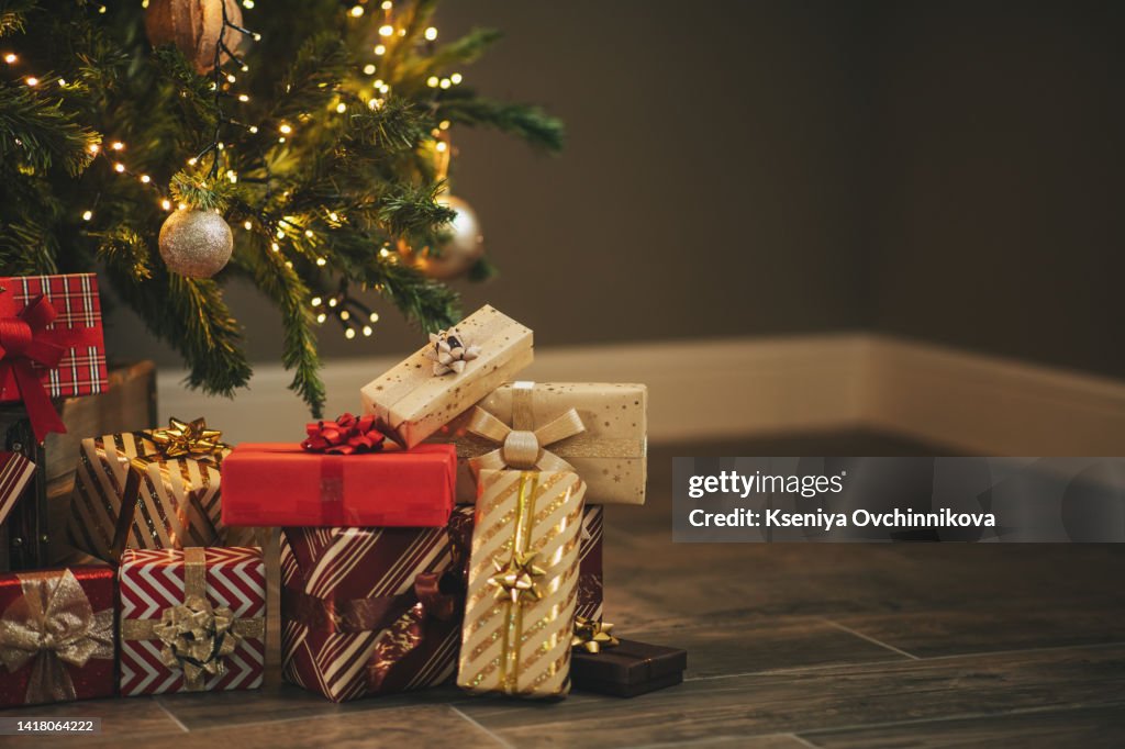 Photo of luxury gift boxes under Christmas tree, New Year home decorations, golden wrapping of Santa presents, festive tree decorated with garland, baubles, traditional celebration. Copy space