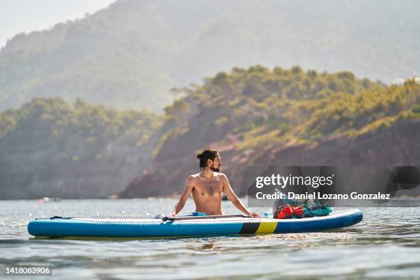 man looking away leaning on his paddle board while relaxing after paddling. - paddle board men imagens e fotografias de stock