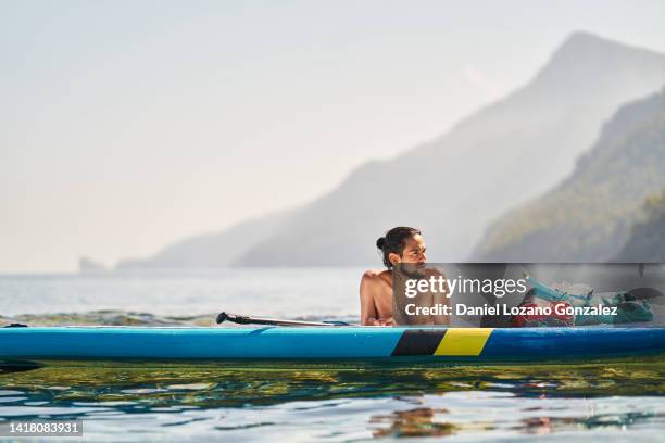 man looking away while relaxing leaning on his paddle surf board in the sea. - paddle board men imagens e fotografias de stock