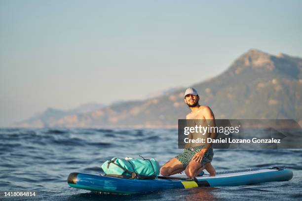 man in cap looking at camera while sitting on paddle board in the sea. - paddle board men imagens e fotografias de stock
