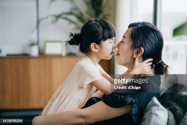 side profile of lovely little asian girl embracing and kissing her mother while relaxing at home. bonding between mother and daughter. family lifestyle. love and care concept - asian mom kid kiss stock-fotos und bilder