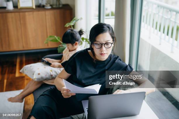 asian mother working from home on laptop while little daughter is watching on digital tablet. asian family using technologies at home. home office and business. handling financial bills with e-banking. working mom managing work life and childcare at home - online bank account stock pictures, royalty-free photos & images