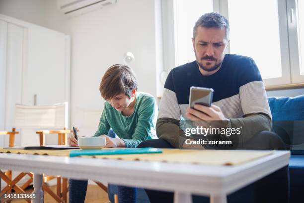 padre ayudando a su hijo educado en casa en la tarea, haciendo investigaciones sobre teléfonos inteligentes - padre soltero fotografías e imágenes de stock