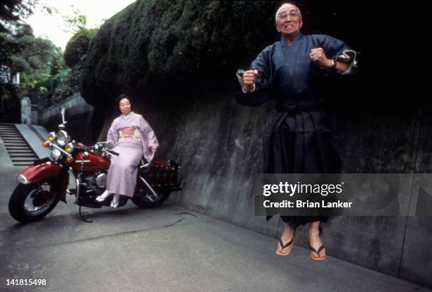 Portrait of Japan Olympian Chuhei Nambu jumping with wife Hisako in background sitting on Harley-Davidson motorcycle. Nambu won the 1932 gold medal...