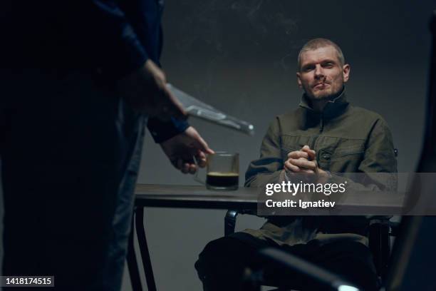 a prisoner in a dark interrogation room sitting in front of an officer, putting a cup of coffee on a table - criminal offense stock pictures, royalty-free photos & images