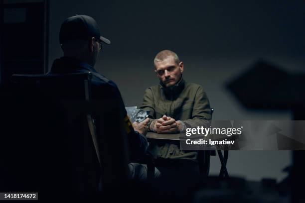 ein gefangener in einem dunklen verhörraum sitzt vor einem beamten mit fotos des verdächtigen - interrogation room stock-fotos und bilder