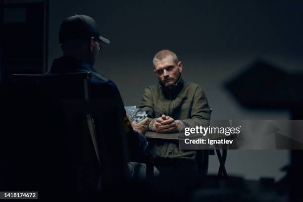 a prisoner in a dark interrogation room sitting in front of an officer with photos of the suspect - interrogated stock pictures, royalty-free photos & images