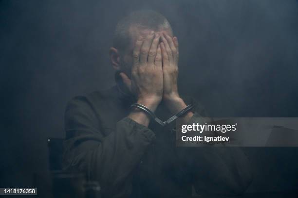 close-up of a suspect in handcuffs closing his face with hands in a dark fogged room - prison uniform stock pictures, royalty-free photos & images