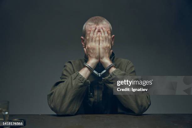 convicto cierra la cara con las manos sobre un fondo gris, sentado en una mesa - sentencing fotografías e imágenes de stock