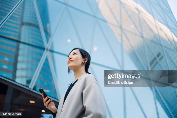 confident young asian businesswoman using smartphone in financial district on the go, standing against urban skyscrapers in the city and looking ahead. successful female entrepreneur looking up to sky. female leadership. business success and achievement - open sky stockfoto's en -beelden