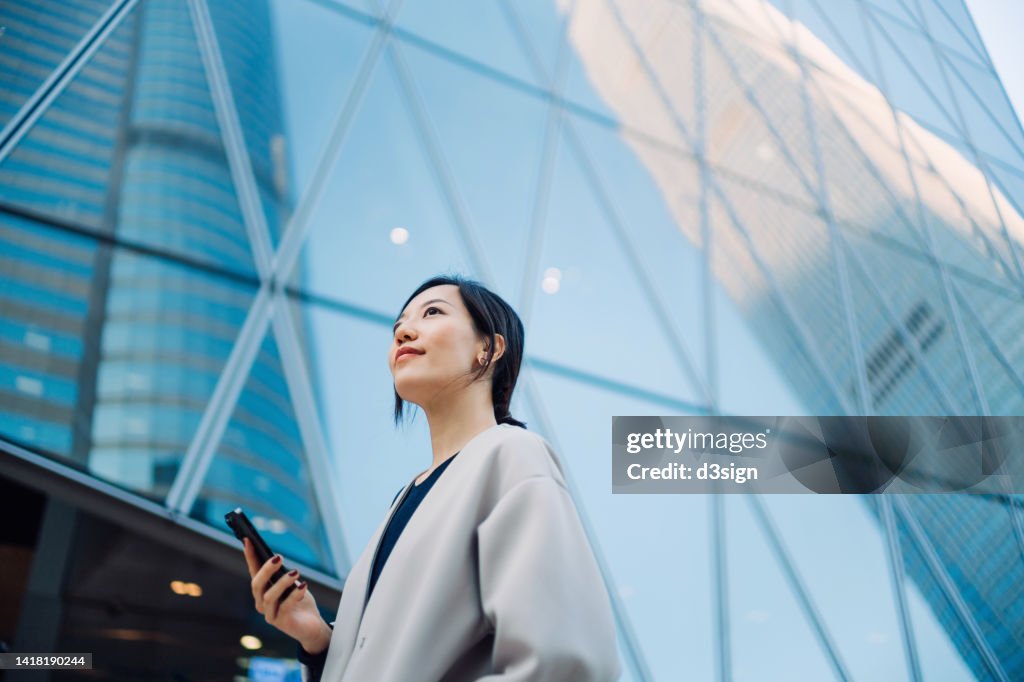 Confident young Asian businesswoman using smartphone in financial district on the go, standing against urban skyscrapers in the city and looking ahead. Successful female entrepreneur looking up to sky. Female leadership. Business success and achievement