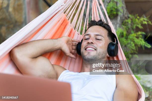 smiling man listening to music and using the computer while lying in a hammock - young man listening music and relaxing in hammock stock pictures, royalty-free photos & images