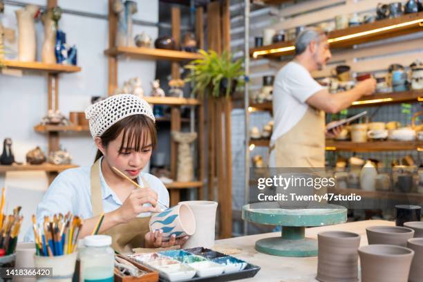 young asian woman potter artist using a paintbrush to paint a ceramic cup in the pottery studio - asian cup draw imagens e fotografias de stock