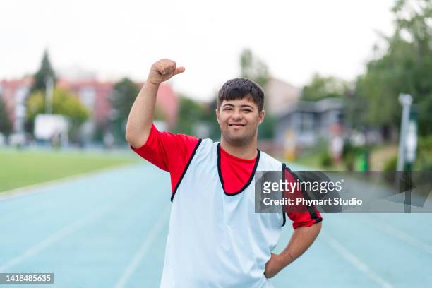 young male athlete with down syndrome posing at sport track and flexing muscles - paralympics track stock pictures, royalty-free photos & images