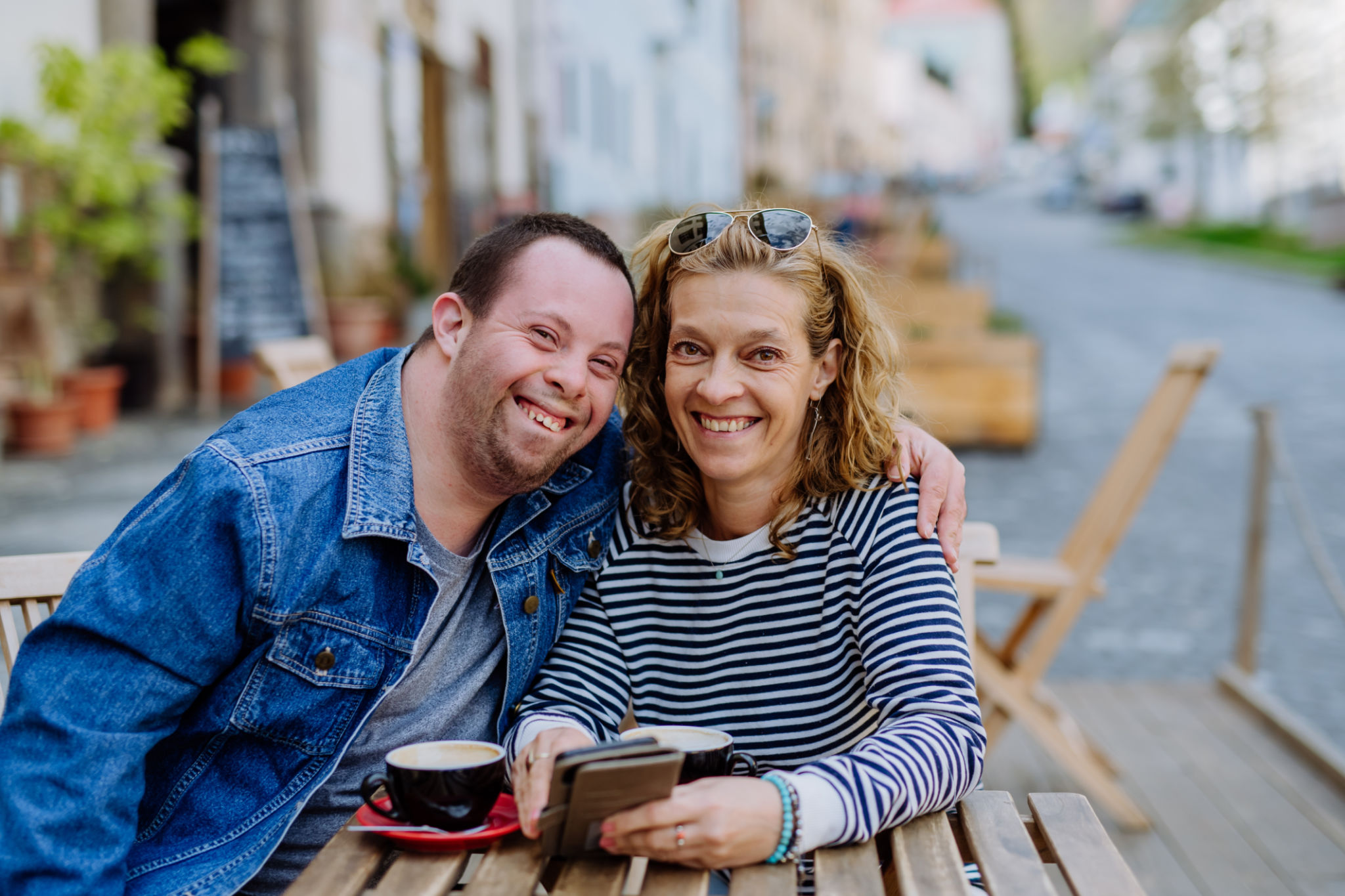 Portrait of happy young man with Down syndrome with his mother sitting at cafe outdoors and talking. Portrait of happy young man with Down syndrome with his mother sitting at cafe outdoors and talking.