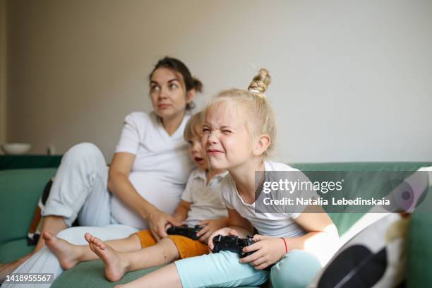 sibling children with pregnant mother sit on sofa and play with joysticks video game together - miopia foto e immagini stock