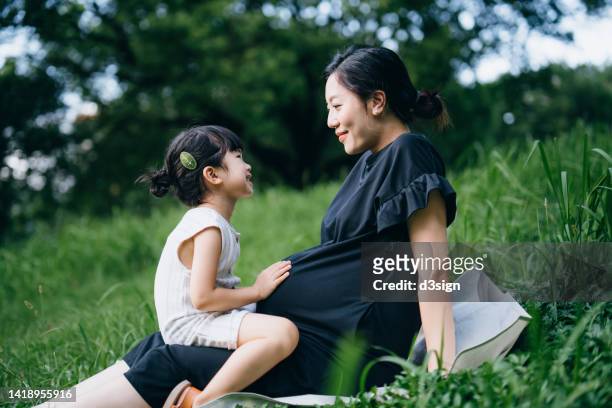 lovely little asian girl gently touching her pregnant mother's baby. big sister talking to the baby and feels the movement of baby in the belly of mother while sitting on meadow in the nature. sibling love. expecting a new life with love and care concept - asian mom kid kiss stock-fotos und bilder