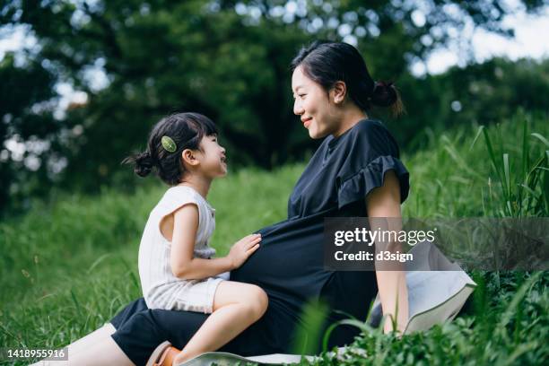 lovely little asian girl gently touching her pregnant mother's baby. big sister talking to the baby and feels the movement of baby in the belly of mother while sitting on meadow in the nature. sibling love. expecting a new life with love and care concept - prenatal care ストックフォトと画像