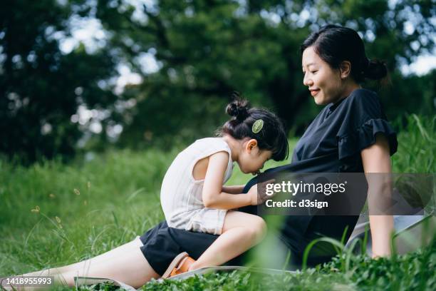 lovely little asian girl gently touching and kissing her pregnant mother's belly while sitting on meadow in the nature. sibling love. expecting a new life with love and care. sharing love concept - belly kissing stock pictures, royalty-free photos & images