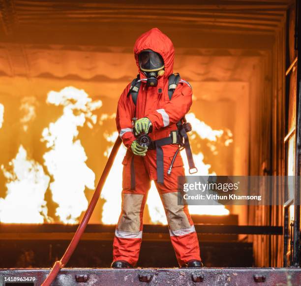 firefighter and protection suit holding fire hose against background burning - roupa a prova de fogo - fotografias e filmes do acervo