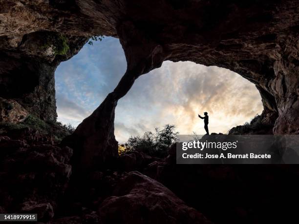 silhouette of a person inside a large rock cave at sunset. - speleology stock pictures, royalty-free photos & images