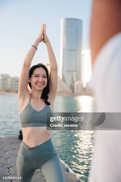 smiling woman doing yoga with her friend by the sea in abu dhabi, arms raised with hands clasped - yoga office arab stock pictures, royalty-free photos & images