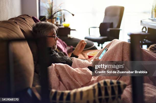 man with gray hair, reading glasses and a few days' beard sitting on the sofa with a blanket over him eating popcorn and looking at the smartphone - covering gray hair stock pictures, royalty-free photos & images