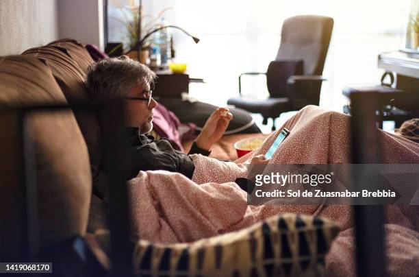 man with gray hair, reading glasses and a few days' beard sitting on the sofa with a blanket over him eating popcorn and looking at the smartphone - persona de color 個照片及圖片檔