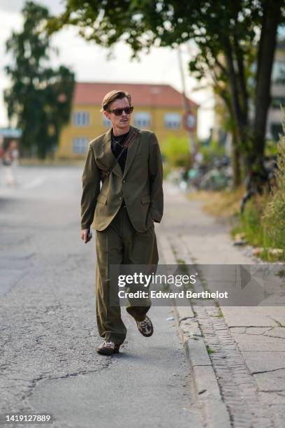 Guest wears brown sunglasses, a black t-shirt, a gold chain pendant necklace, a khaki blazer jacket, matching khaki large suit pants, a brown leather...