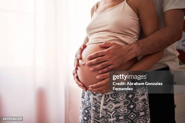 man touching pregnant woman's stomach while standing in room near window - husband stock pictures, royalty-free photos & images