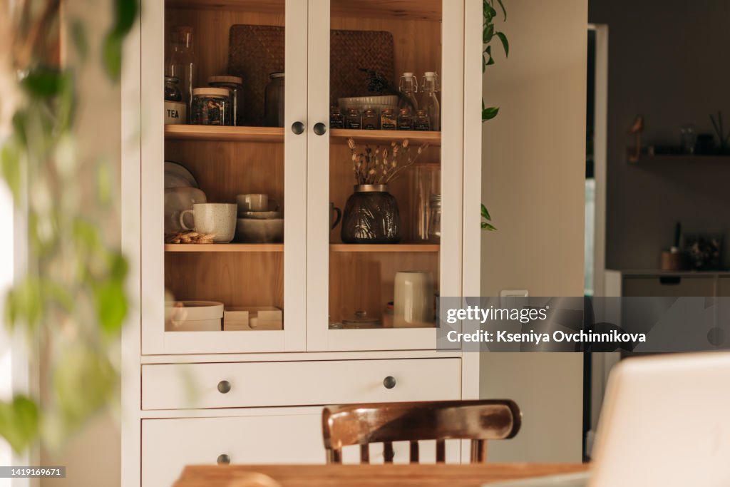 Opened white glass cabinet with clean dishes and decor. Scandinavian style kitchen interior. Organization of storage in kitchen.