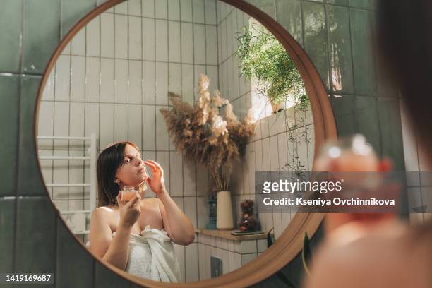 young brunette girl taking care of her skin, standing in front of mirror with eyes closed, enjoying beauty treatment for herself, smiling gently - woman in bathroom stockfoto's en -beelden
