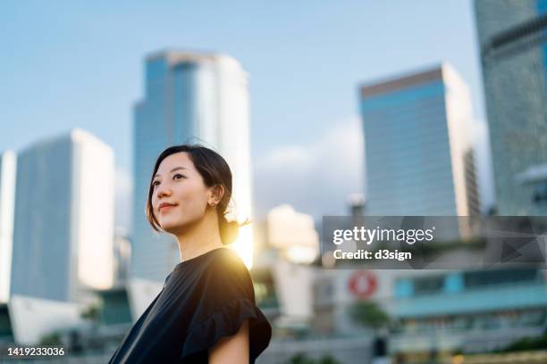 confident and ambitious young asian businesswoman looking ahead with positive emotion, standing against prosperous urban city skyline in downtown district. girl power. successful female entrepreneur at work. female leadership and determined to success - corporate gender equality stock pictures, royalty-free photos & images