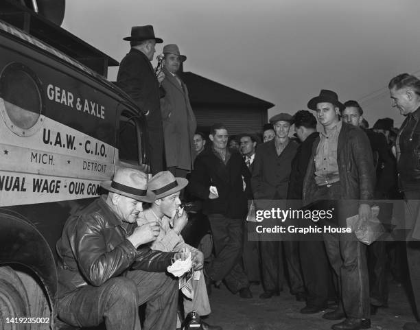 Man in an overcoat and fedora addressing a crowd of General Motors employees, as two men eat while sitting on the running board of a vehicle outside...