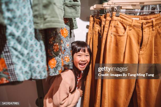 lovely little girl smiling joyfully behind a clothes rack while shopping in a clothing store - local girls fotografías e imágenes de stock