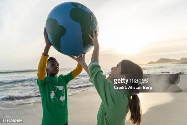 young activists holding globe up on a beach at sunset - global crisis stock pictures, royalty-free photos & images