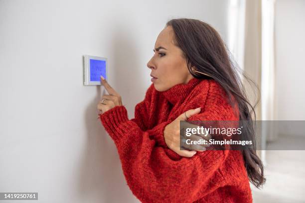 freezing woman at home wears sweater and tries to raise the temperature on thermostat while energy crisis hits europe in the winter. - electric heater fotografías e imágenes de stock