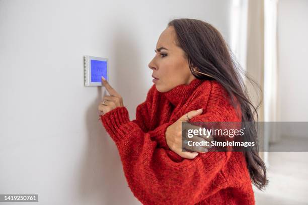 freezing woman at home wears sweater and tries to raise the temperature on thermostat while energy crisis hits europe in the winter. - cold temperature foto e immagini stock