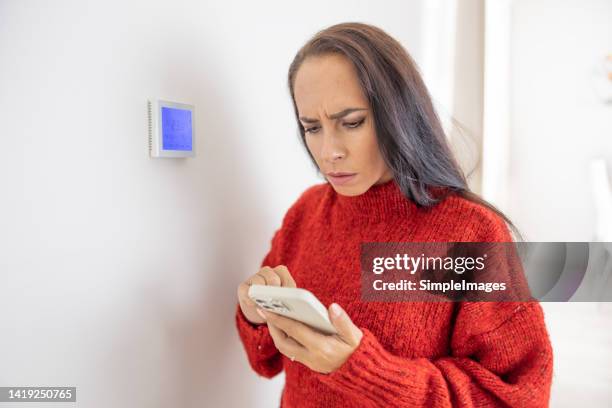 woman in red sweater tries to lower the temperature on the heating at home via smartphone application. - low temperature stock pictures, royalty-free photos & images