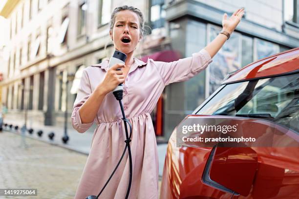 portrait of young woman charging her electric car and having fun - singing microphone stock pictures, royalty-free photos & images