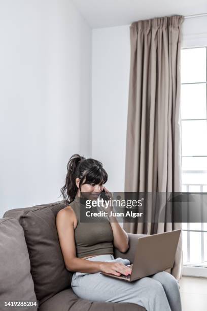 smiling woman sitting on the sofa at home talking on the phone with a laptop on her lap - habitación stockfoto's en -beelden